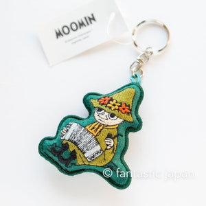May include: Green felt keychain featuring an embroidered Moomin character playing an accordion. The character is wearing a green hat with flowers and a yellow scarf. The keychain has a silver metal ring.