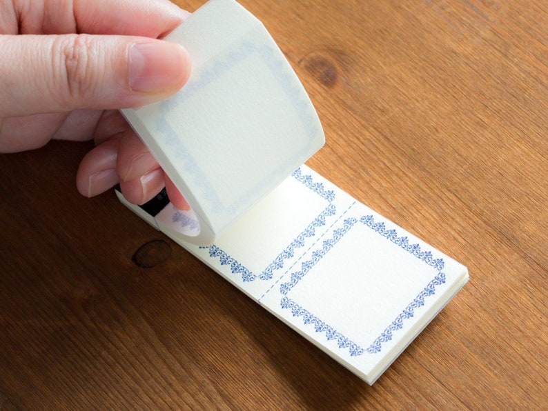 Puede incluir: Un rollo de cinta washi decorativa con un dise&ntilde;o de borde azul y blanco. La cinta se est&aacute; desenrollando para revelar una hoja de papel blanco con dos marcos de borde azul y blanco.