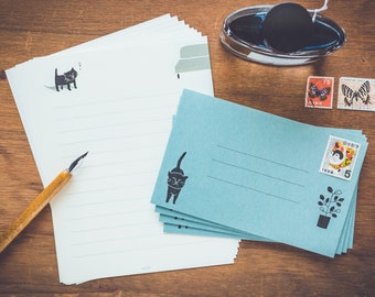 Puede incluir: Una pila de papel de cartas azul con una ilustración de un gato negro y una nota manuscrita. El papel está sobre una superficie de madera con un bolígrafo, un tintero y sellos.