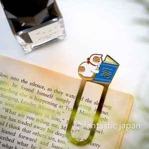 May include: Gold bookmark featuring a white cat with a red bow reading a blue book with Japanese text. The bookmark is shaped like a cat's tail.