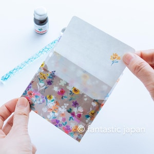 May include: A floral patterned stationery set featuring a translucent envelope with a colorful flower design. The set includes a lined writing sheet with a small yellow flower illustration, a blue pen, and a bottle of ink.
