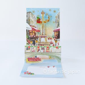 May include: A pop-up Christmas card featuring a festive winter scene. The card depicts a snowy street with shops, a bridge, and a tower. Santa Claus flies in a sleigh pulled by reindeer. The card has a blue background with snowflakes.