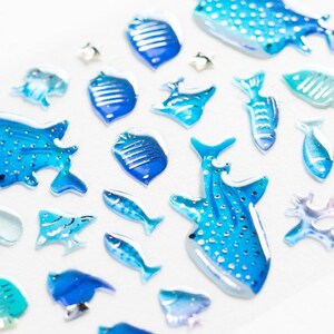 May include: A collection of blue and white resin stickers featuring various sea creatures, including whale sharks, fish, and coral. The stickers are ideal for adding a touch of ocean charm to crafts, journals, and more.
