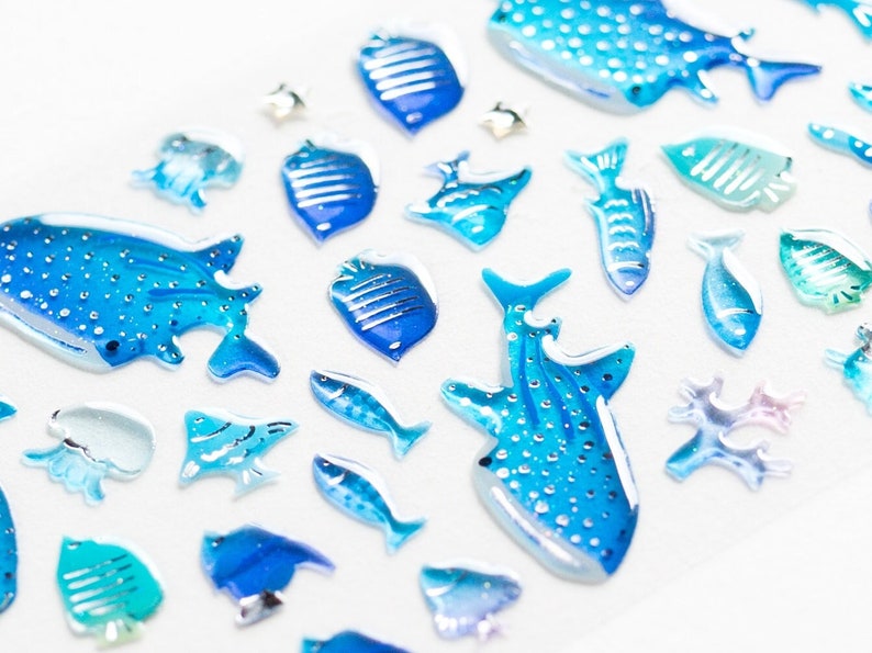 May include: A collection of blue and white resin stickers featuring various sea creatures, including whale sharks, fish, and coral. The stickers are ideal for adding a touch of ocean charm to crafts, journals, and more.