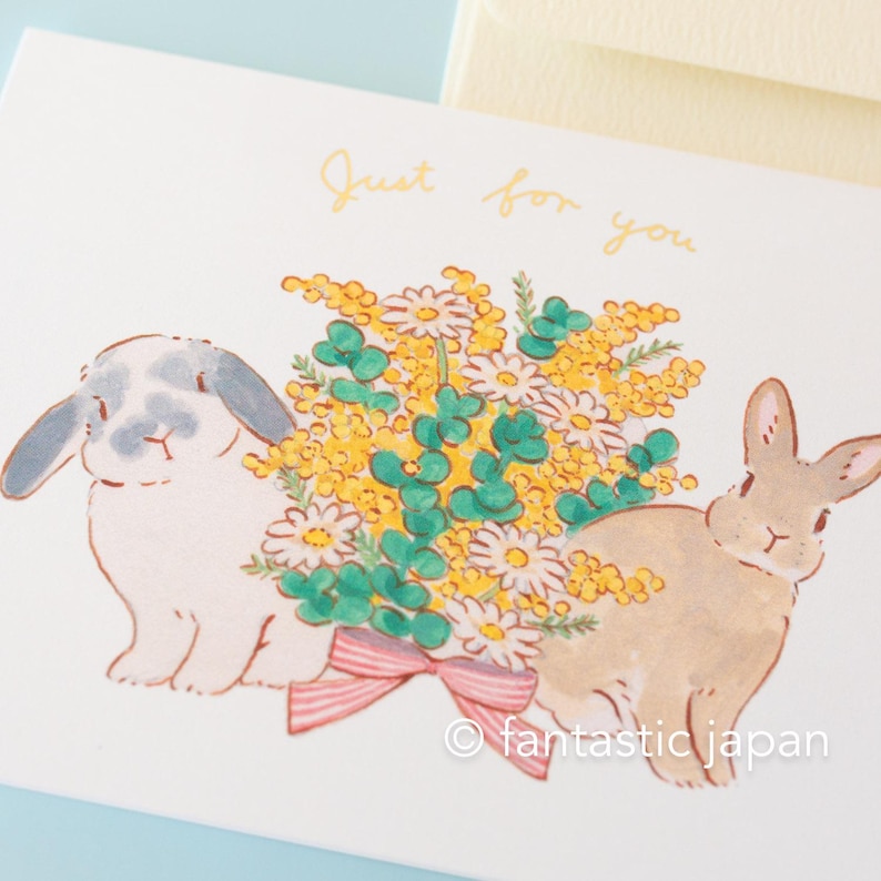 May include: A greeting card featuring two bunnies and a bouquet of yellow and white flowers. The card reads "Just for you" in yellow script. The bunnies are holding a pink ribbon.
