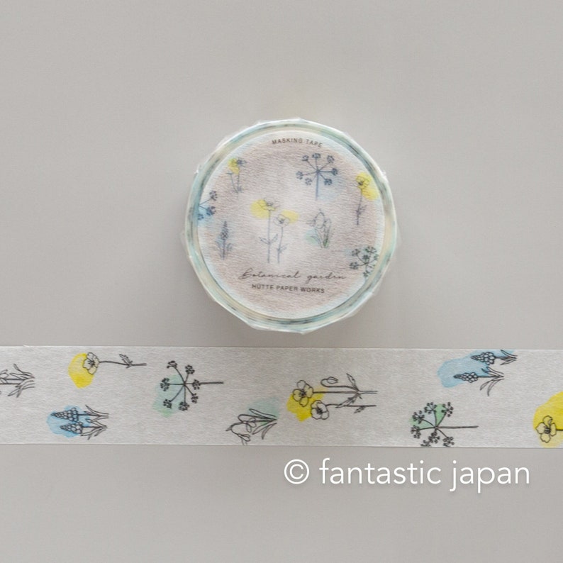 Puede incluir: Un rollo de cinta washi con un dise&ntilde;o floral. La cinta es blanca con acentos acuarela azul y amarillo. La cinta est&aacute; etiquetada como "Botanical Garden" y "Mutte Paper Works".