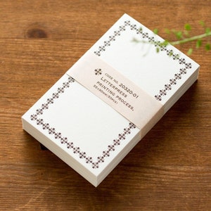 Könnte beinhalten: Ein Stapel Briefpapier mit floralem Bordürendesign. Das cremefarbene Papier ist mit einem Band mit dem Text "CODE NO. 20320-01 LETTERPRESS PRINTING PROCESS, 55x90mm (50PCS)" umwickelt. Das Briefpapier liegt auf einer Holzoberfläche.