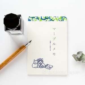 Könnte beinhalten: Ein weißer Notizblock mit einem blauen und grünen Muster an der Kante. Der Notizblock trägt den Text "マールメ" in Japanisch, was "Marlme" bedeutet. Der Notizblock trägt auch den Text "素敵文具専用" in Japanisch, was "Exklusiv für schöne Schreibwaren" bedeutet. Der Notizblock zeigt eine blaue Illustration von zwei Katzen, eine davon mit einem Hut, die neben einem Buch sitzen. Der Notizblock liegt auf einer weißen Oberfläche mit einem schwarzen Tintenfass und einem Holzstift.