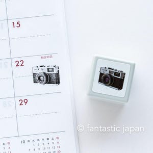 May include: A gray rubber stamp with a black and white illustration of a vintage camera. The stamp is on a white background with a calendar page behind it. The calendar page has the text "秋分の日" which translates to "Autumn Equinox" in English.