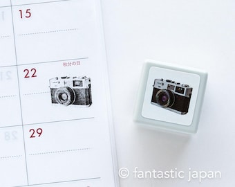 May include: A grey rubber stamp featuring a black and white illustration of a vintage camera. The stamp is on a white background with a calendar page behind it. The calendar page has the text "秋分の日" which translates to "Autumn Equinox" in English.