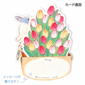 May include: A pop-up card featuring a white rabbit with blue accents peeking out from behind a bouquet of pink, yellow, and white tulips. The card has a blank space for a message and the text "Message" printed above it. The card is made of paper and has a wooden base.