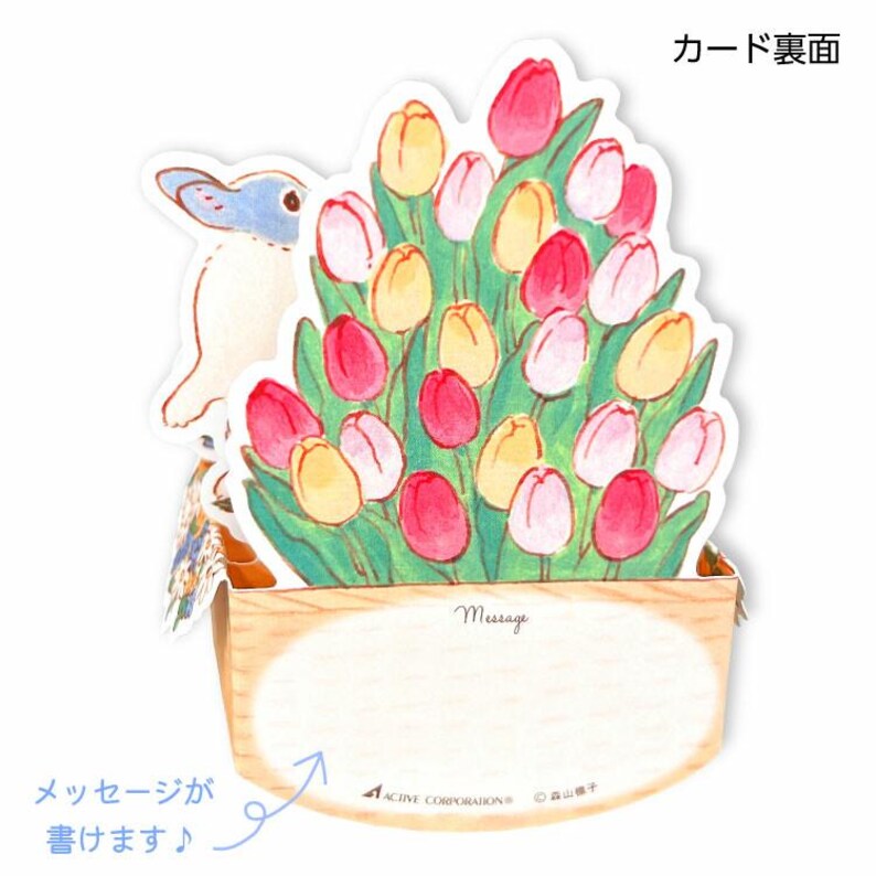 May include: A pop-up card featuring a white rabbit with blue accents peeking out from behind a bouquet of pink, yellow, and white tulips. The card has a blank space for a message and the text "Message" printed above it. The card is made of paper and has a wooden base.