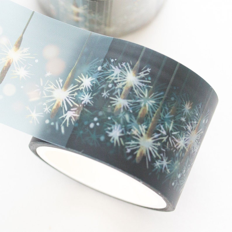 May include: A roll of decorative washi tape featuring a dark background with white and light blue starburst-like designs. The tape has a white interior and is partially unrolled, revealing the design.