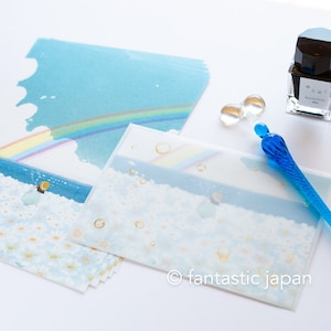 May include: Stationery set featuring a blue and white design with a rainbow and daisy field illustration. Includes envelopes, a blue glass pen, and a bottle of ink. The set has a whimsical, artistic aesthetic.
