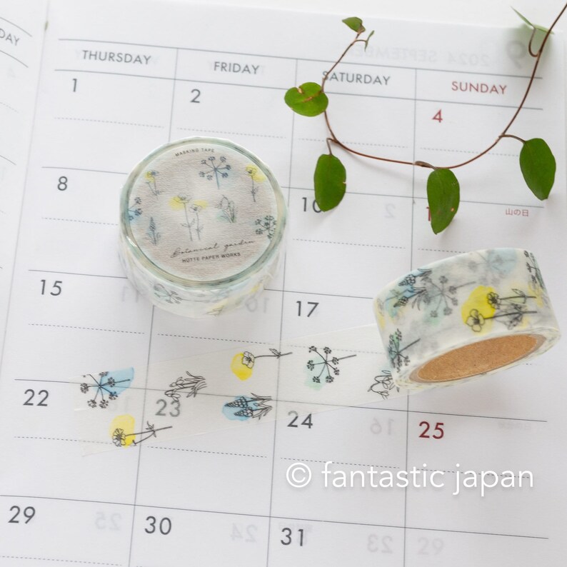 Puede incluir: Dos rollos de cinta washi con un patr&oacute;n floral. La cinta es blanca con flores y hojas verdes y amarillas. El texto en la cinta dice "Botanical Garden" y "Motif Paper Works".