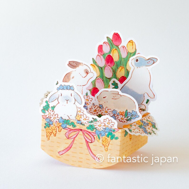 May include: A pop-up card featuring a basket filled with flowers and four bunnies. The basket is yellow with a pink ribbon. The flowers are pink, yellow, and white. The bunnies are white, gray, and brown. The card says "fantastic japan" at the bottom.