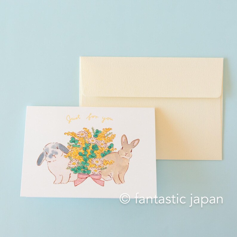 May include: A white card with a hand-drawn illustration of two rabbits with a bouquet of yellow and white flowers. The text "Just for you" is written in gold lettering on the card.