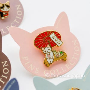 May include: A gold-colored enamel pin featuring a white cat under a red parasol, with the Japanese word "dango" on a banner. The cat is sitting on a wooden bench with dango and tea. The pin is on a pink cat-shaped backing.