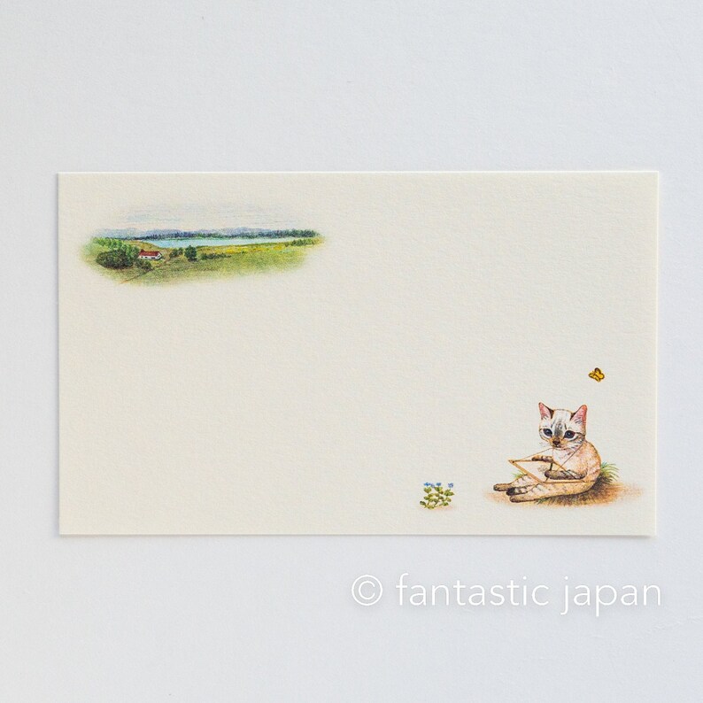 May include: A cream-colored stationery card featuring a watercolor illustration. The card depicts a landscape scene with a house, trees, and a lake. A cat is sitting on the grass, holding a paper, with a butterfly nearby. The text "fantastic japan" is at the bottom.