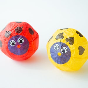 Puede incluir: Dos farolillos de papel con forma de mariquita. Uno es rojo con manchas negras y el otro es amarillo con corazones negros. Ambos tienen caras azules con ojos negros y una nariz roja.
