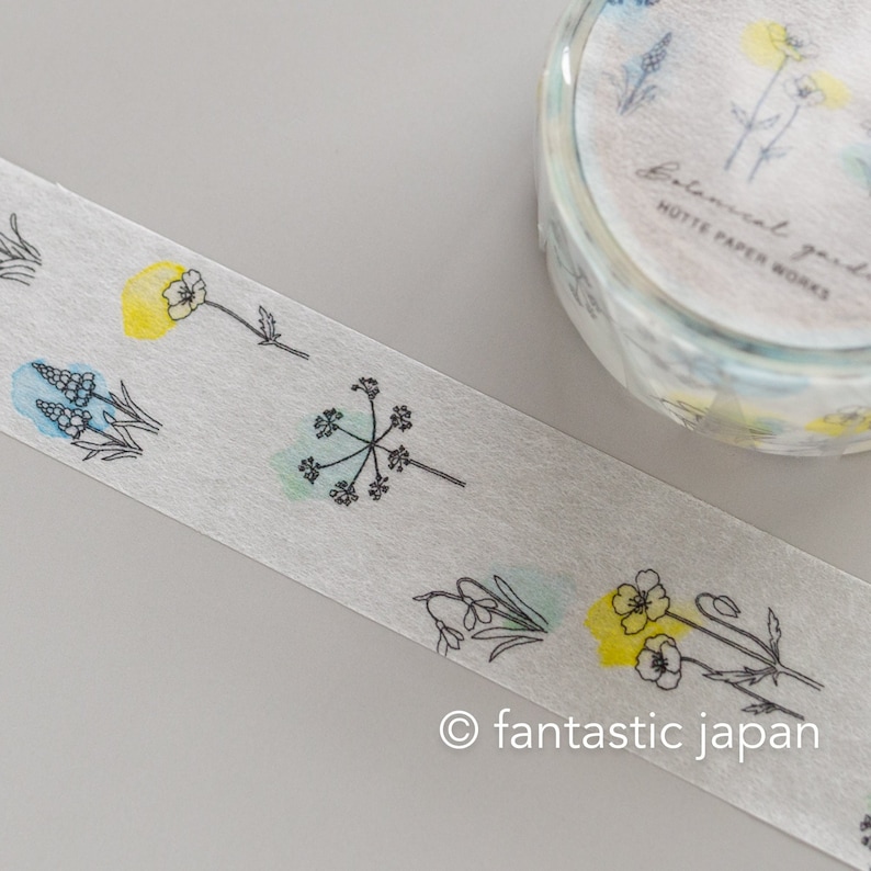 Puede incluir: Un rollo de cinta washi con un dise&ntilde;o floral. La cinta presenta delicados dibujos de l&iacute;nea negra de flores y hojas con lavados de acuarela en tonos azules, verdes y amarillos. El texto "Botanical garden" y "Hutte Paper Works" est&aacute; impreso en la cinta.