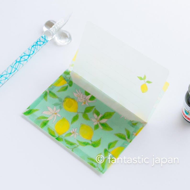 May include: An open stationery set featuring a lemon design. The set includes a notepad with lined pages and a matching envelope, decorated with yellow lemons, white blossoms, and green leaves on a light green background. A glass pen with blue accents is visible.