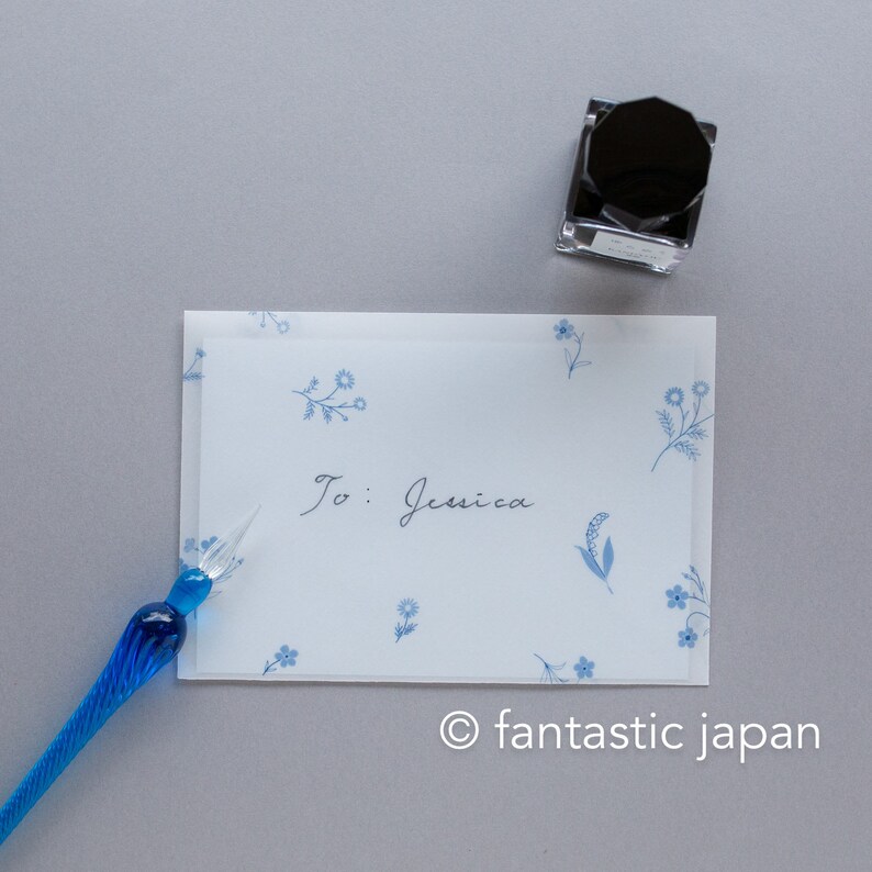 Pode incluir: Uma caneta de vidro azul est&aacute; ao lado de um cart&atilde;o com o texto "To: Jessica" e desenhos florais azuis. Um frasco de tinta preta est&aacute; por perto. O cart&atilde;o e a caneta est&atilde;o sobre uma superf&iacute;cie cinza.