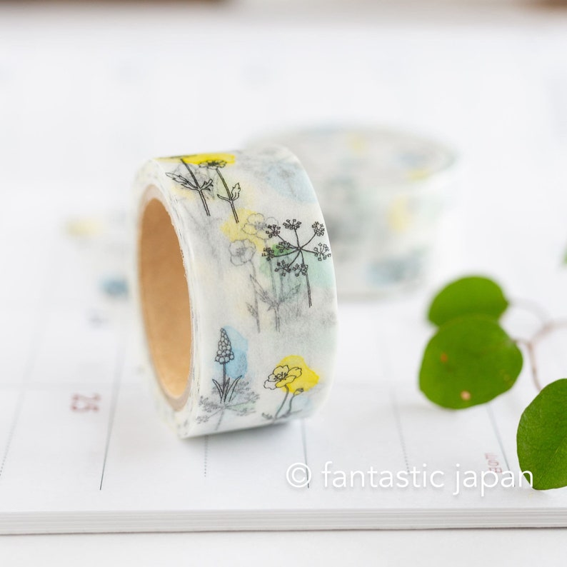 Puede incluir: Un rollo de cinta washi con un dise&ntilde;o floral que presenta flores de acuarela amarillas, azules y verdes y contornos negros sobre un fondo blanco.
