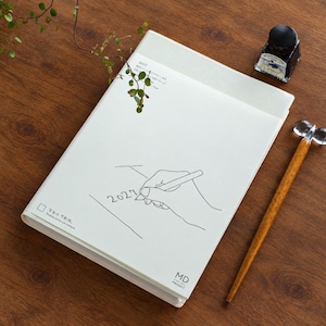 Könnte beinhalten: Ein weißes MD-Notizbuch mit einem 2027-Cover-Grafikdesign, das eine Strichzeichnung einer schreibenden Hand, den Text 2027 und den Satz Passing notes you forward zeigt. Das Notizbuch wird von einem Glastintenfläschchen und einem Holzstift begleitet.