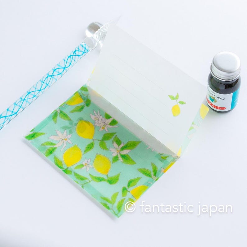May include: A stationery set featuring a lemon theme. The set includes a notepad with lined pages and a lemon illustration, a glass pen with a blue design, and a bottle of ink. The notepad has a lemon and floral pattern on the cover.