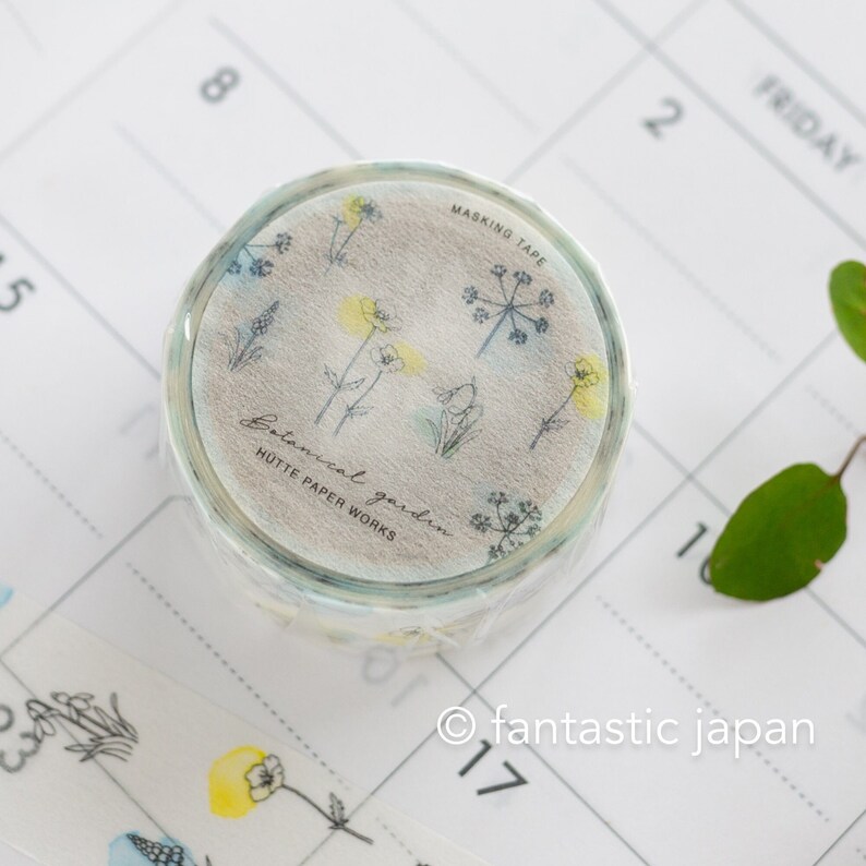Puede incluir: Un rollo de cinta washi con un dise&ntilde;o floral. La cinta es blanca con un fondo acuarela azul claro y presenta delicados dibujos en negro de flores y plantas. El texto "Botanical garden" y "H&Uuml;TTE PAPER WORKS" est&aacute; impreso en la cinta.
