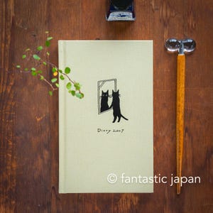 Könnte beinhalten: Ein hellgrünes Hardcover-Tagebuch mit dem Text Diary 2027 und einer schwarzen Illustration einer Katze, die in einen Spiegel schaut. Neben dem Tagebuch liegt ein hölzerner Tauchfüller mit einem klaren Glasgriff und ein kleines Glas mit dunkelblauer Tinte.