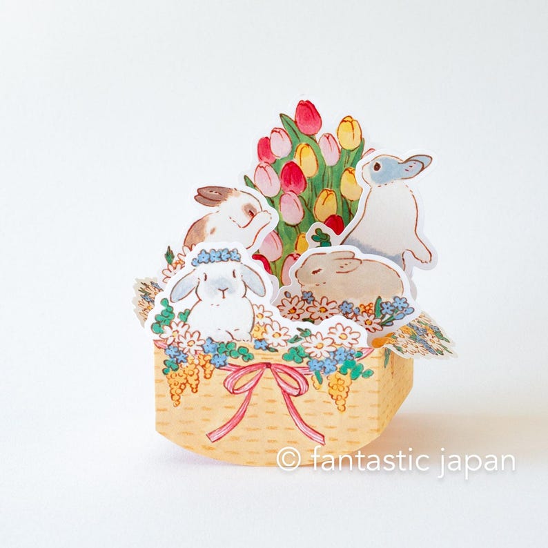 May include: A pop-up card featuring a basket filled with flowers and four bunnies. The basket is decorated with a pink ribbon and the flowers are a mix of pink, yellow, and white. The bunnies are all white with pink noses and whiskers. The card is made of paper and has a white background.