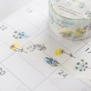 Puede incluir: Un rollo de cinta washi con un dise&ntilde;o floral. La cinta presenta delicados dibujos de flores y hojas en l&iacute;nea negra con lavados de acuarela en tonos azules, amarillos y verdes. La cinta est&aacute; etiquetada como "Botanical Garden Hutte Paper Works".