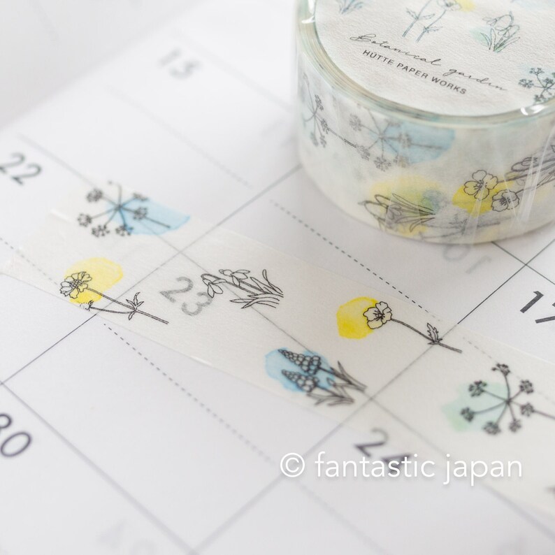 Puede incluir: Un rollo de cinta washi con un dise&ntilde;o floral. La cinta presenta delicados dibujos de flores y hojas en l&iacute;nea negra con lavados de acuarela en tonos azules, amarillos y verdes. La cinta est&aacute; etiquetada como "Botanical Garden Hutte Paper Works".