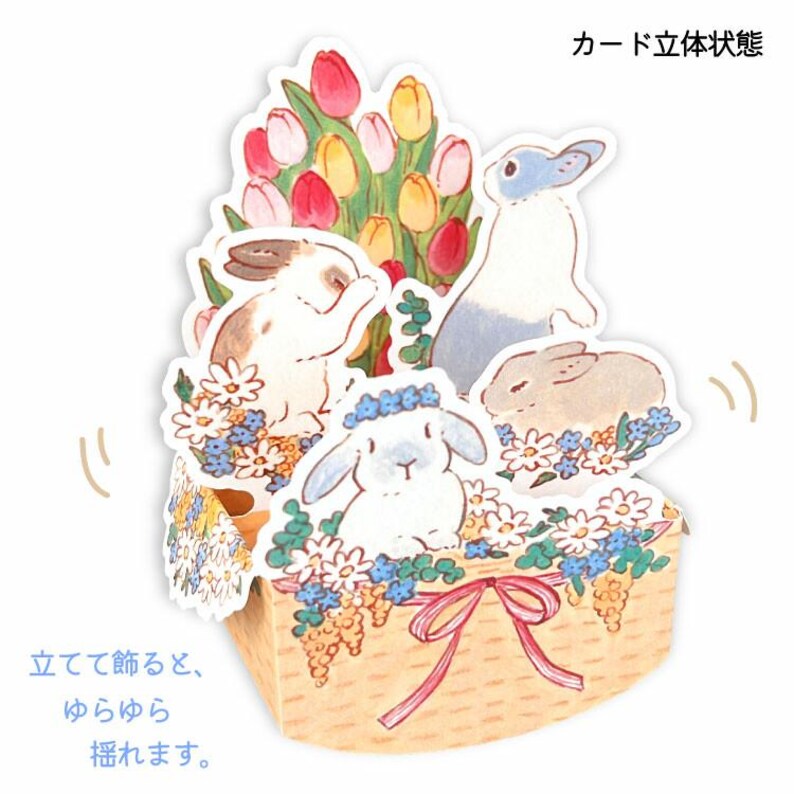 May include: A pop-up card with a basket of flowers and three bunnies. The card is made of paper and has a 3D effect. The text on the card reads "Standing up, the bunnies pop up."