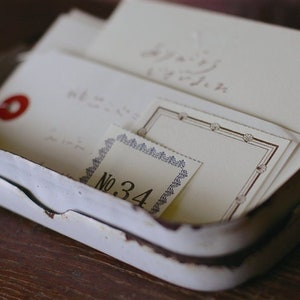 Puede incluir: Una lata de metal blanca con un acabado desgastado y envejecido. La lata contiene varios trozos de papel con texto escrito a mano y bordes decorativos. Un trozo de papel tiene el n&uacute;mero "No. 34" impreso en &eacute;l.