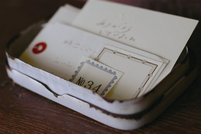 Puede incluir: Una lata de metal blanca con un acabado desgastado y envejecido. La lata contiene varios trozos de papel con texto escrito a mano y bordes decorativos. Un trozo de papel tiene el n&uacute;mero "No. 34" impreso en &eacute;l.