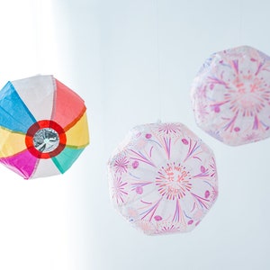 Puede incluir: Tres linternas de papel de colores colgando del techo. Las linternas son redondas y tienen diferentes patrones. Una linterna es multicolor con un círculo plateado en el centro. Las otras dos linternas son blancas con diseños florales rosas y morados.