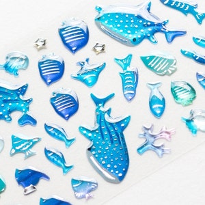 May include: A sheet of blue and white whale shark stickers. The stickers are in various sizes and shapes, including whale sharks, fish, and coral. The stickers are ideal for adding a touch of whimsy to any project.