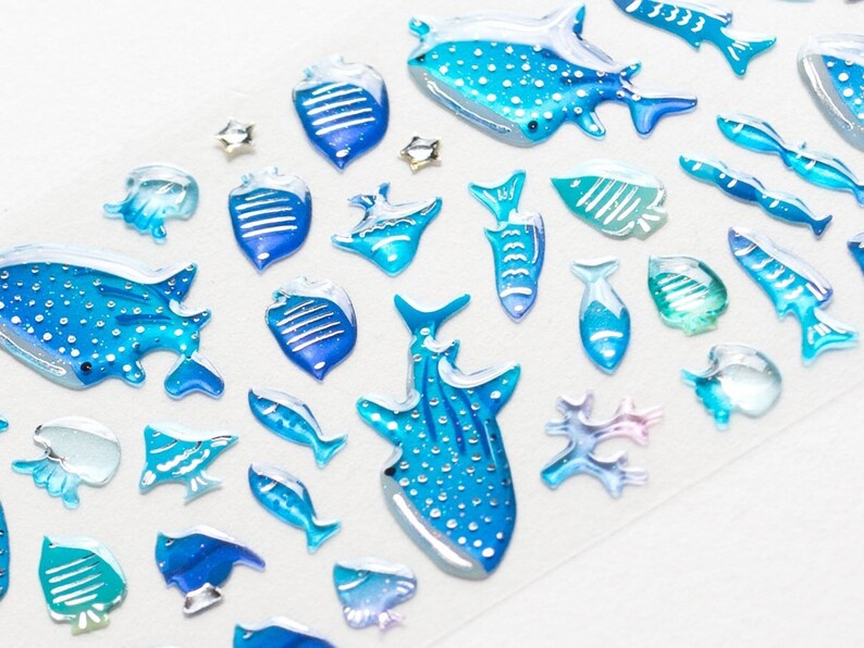 May include: A sheet of blue and white whale shark stickers. The stickers are in various sizes and shapes, including whale sharks, fish, and coral. The stickers are ideal for adding a touch of whimsy to any project.