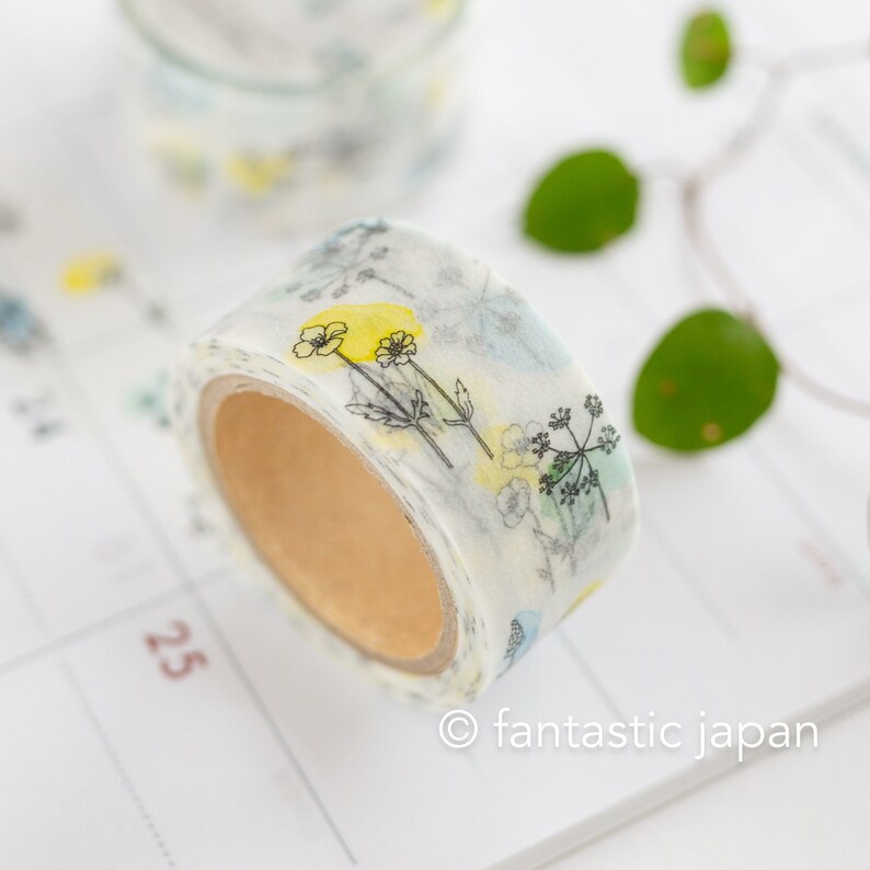 Puede incluir: Un rollo de cinta washi blanca con un dise&ntilde;o floral que presenta flores amarillas y hojas verdes. La cinta est&aacute; sobre una superficie blanca con el texto "fantastic japan" impreso en ella.