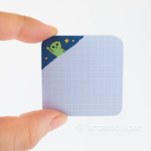 Può includere: Un blocco note a griglia blu chiaro con una linguetta d'angolo che presenta un personaggio alieno verde con stelle su uno sfondo blu scuro.