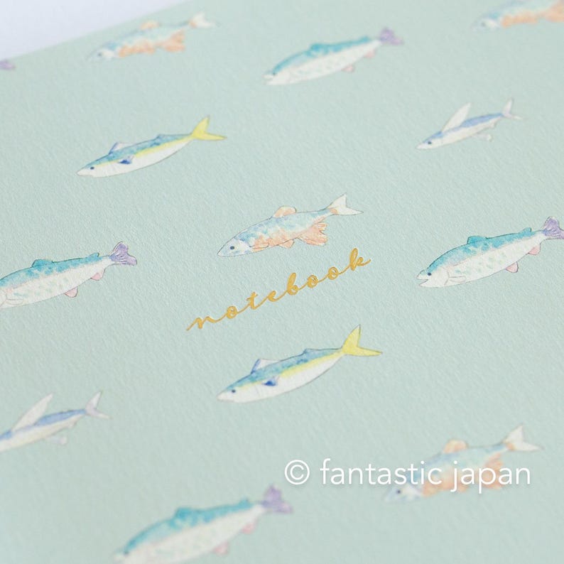 May include: A light blue notebook with a repeating pattern of watercolor fish in shades of blue, yellow, and orange. The word "notebook" is written in gold script. The copyright symbol and the words "fantastic japan" are at the bottom.