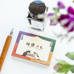 Könnte beinhalten: Ein japanisches Schreibwarenset mit einem Notizblock mit Katzenillustrationen und japanischem Text, einem Glas-Tintenfass und einer Kalligraphie-Feder aus Holz. Der Notizblock hat einen farbenfrohen Rand und den Text "SUMO MEMO".
