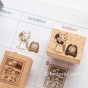 May include: Wooden stamps with cat illustrations. One stamp features a cat on the phone next to a rotary phone. Another stamp shows a cat on a stamp with the number 25. The stamps are on a calendar page.