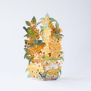 May include: A 3D paper art piece featuring a basket overflowing with yellow flowers and green leaves. Two green birds perch among the flowers. A small sign reads "For you". The artwork is set against a white background.