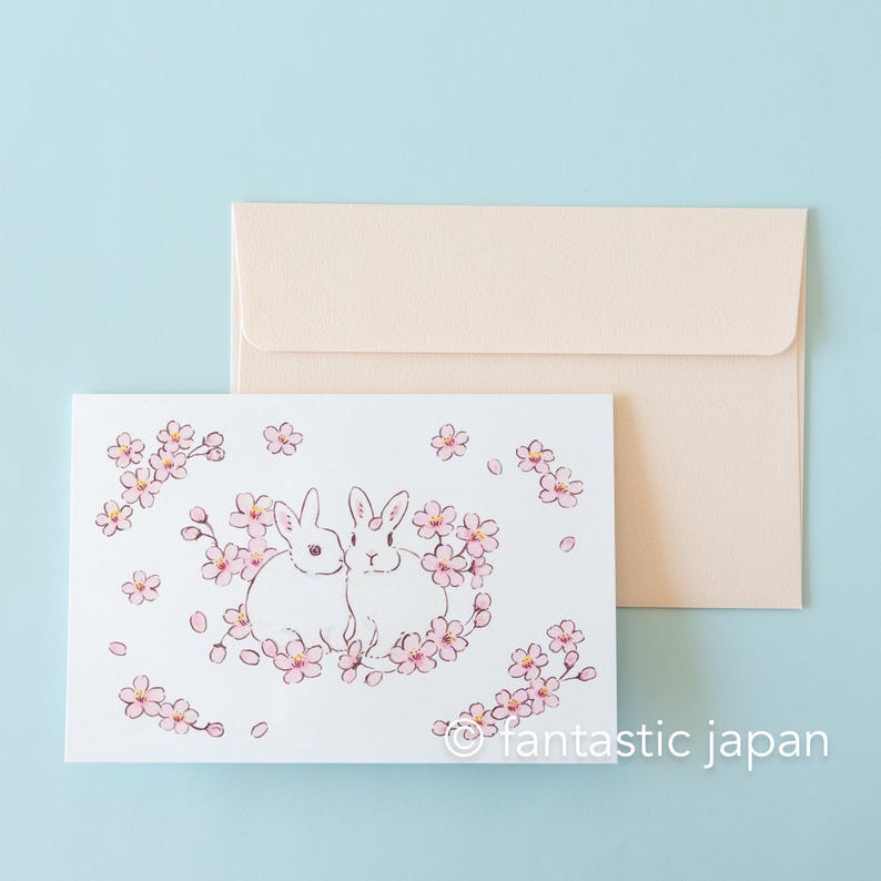 Może przedstawiać: Biała kartka z dwoma białymi kr&oacute;likami siedzącymi w k&oacute;łku r&oacute;żowych kwiat&oacute;w wiśni. Kartka ma białe tło i napis "&copy;fantastic japan" w prawym dolnym rogu.