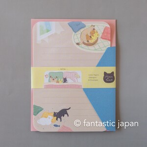 May include: A stationery set featuring illustrated cats. The set includes letter paper with a pink border and a blue envelope. The letter paper has a beige background with cat illustrations and lines for writing. The set is wrapped with a yellow band that has text and cat illustrations.