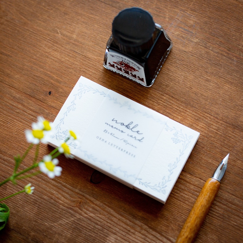 May include: A pack of white memo cards with a blue floral border and the text "noble memo card 8.5 x 6.0cm / Japan O.R.D.A LETTERPRESS" printed on the front. A bottle of black ink and a wooden calligraphy pen are also in the image.